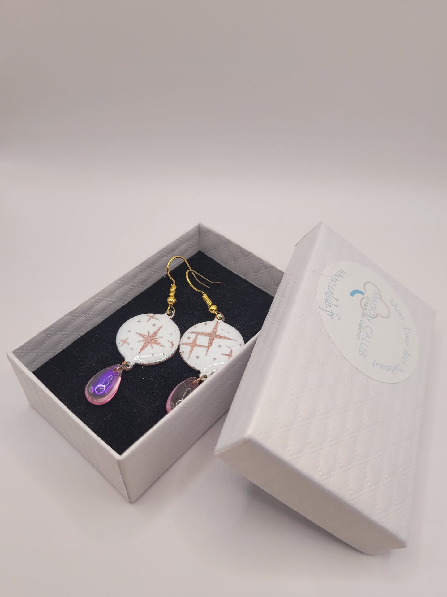 dans leur boite cadeau des boucles d'oreilles en émail sur cuivre aux motif d'étoiles rosés sur blanc prolongé par une perle de verre goutte. Bijou artisanal fait à la main chez maison calub les emaux du Luberon