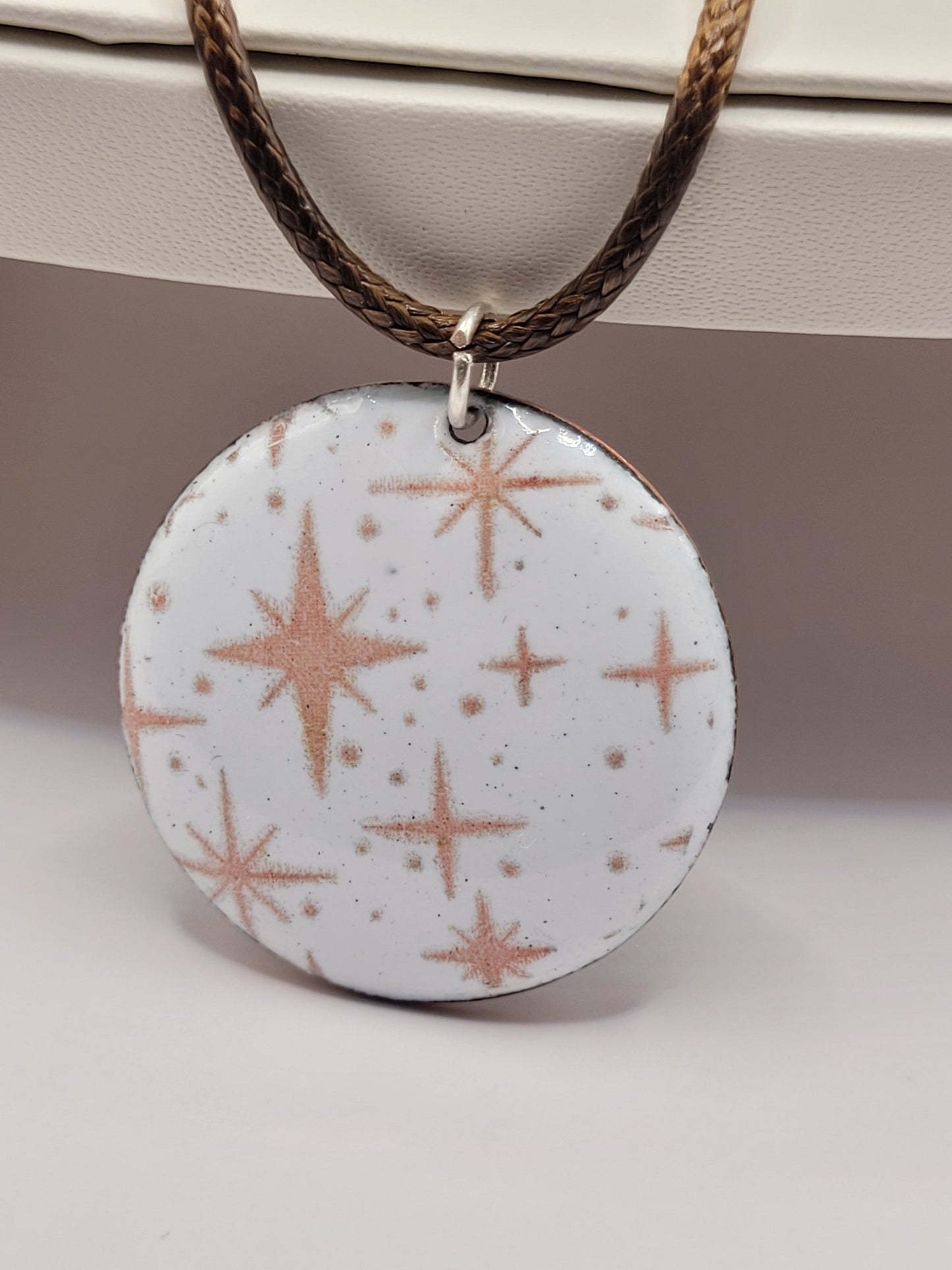 pendentif émaillé lumineux aux fines étoiles rosées sur fond blanc émaux sur cuivre artisanaux de maison calub sur présentoir