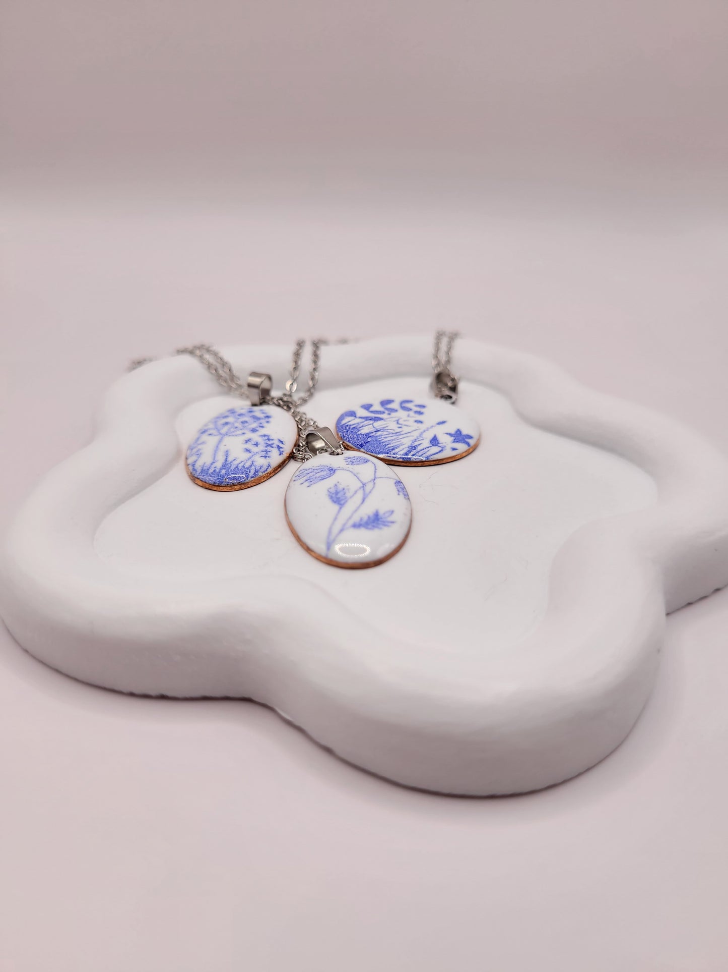 3 pendentifs de la série porcelaine chez maison Calub. Pendentif ovale ou rond en émail sur cuivre blanc porcelaine orné de fins motif floraux bleus, monté sur une fine chaine en acier.