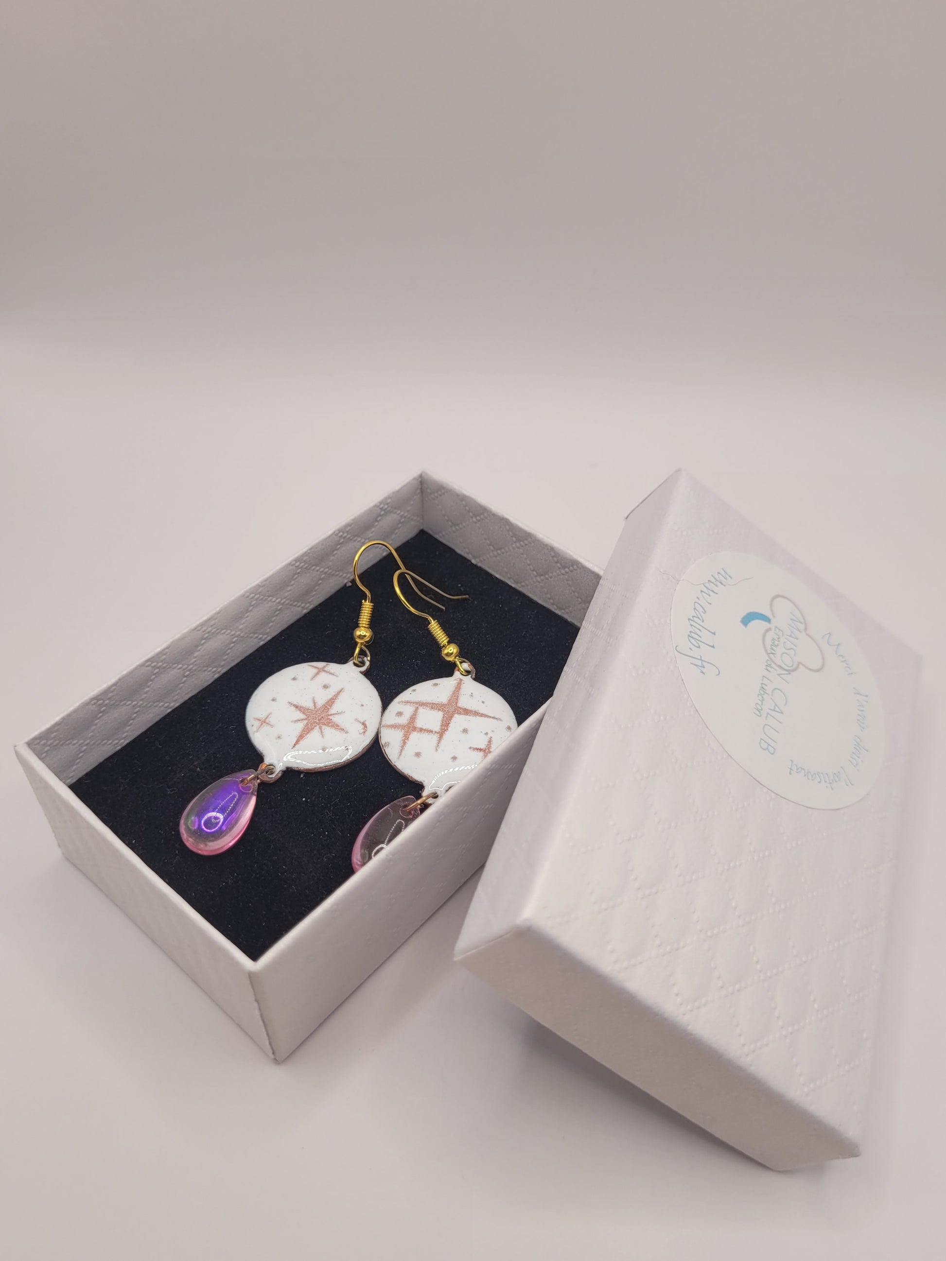 dans leur boite cadeau des boucles d'oreilles en émail sur cuivre aux motif d'étoiles rosés sur blanc prolongé par une perle de verre goutte. Bijou artisanal fait à la main chez maison calub les emaux du Luberon